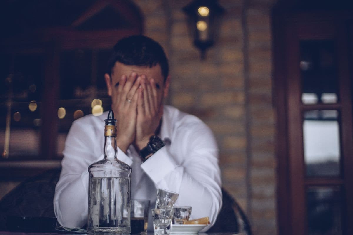 uomo con le mani sul volto e bottiglia vuota di alcol sul tavolo